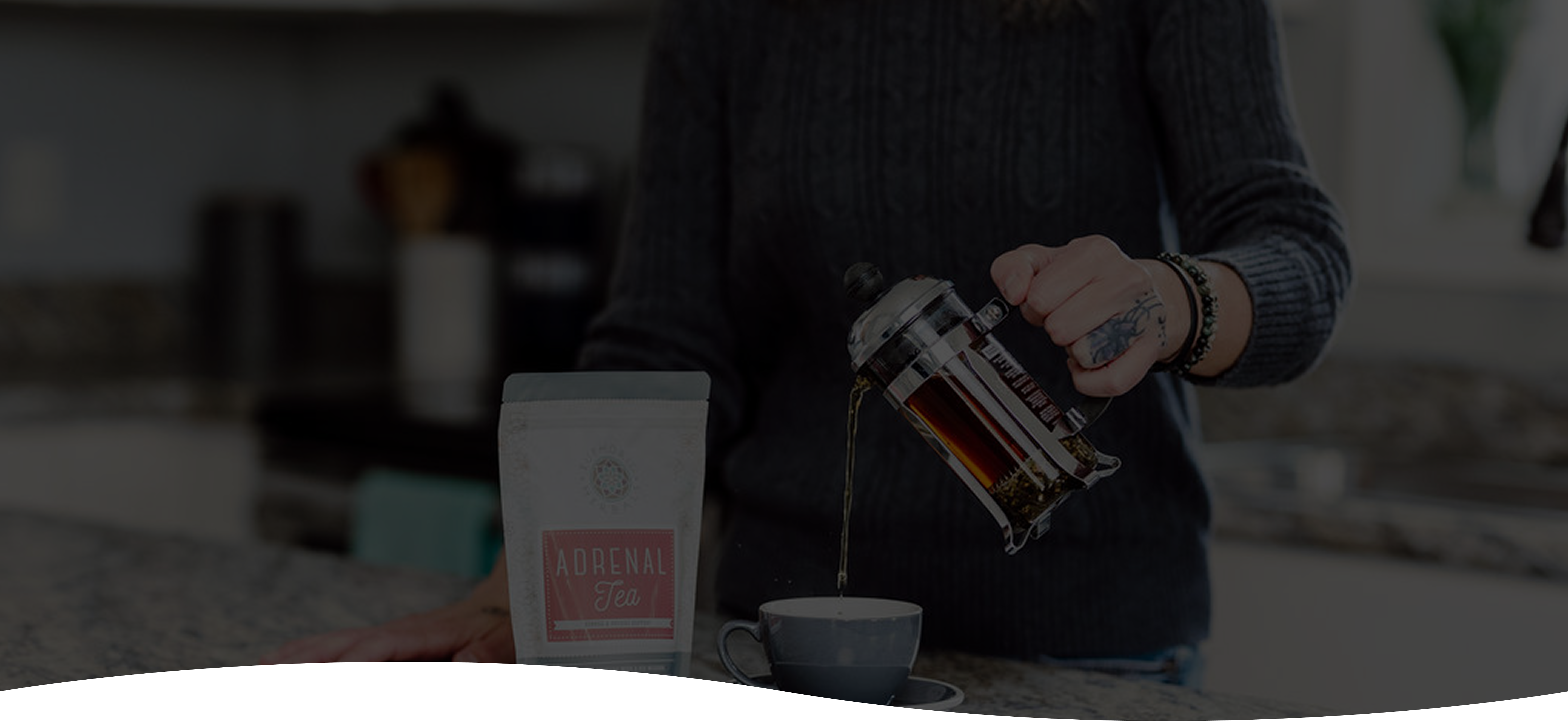 Person pouring tea from a French press into a cup with a bag of herbal tea in the foreground.