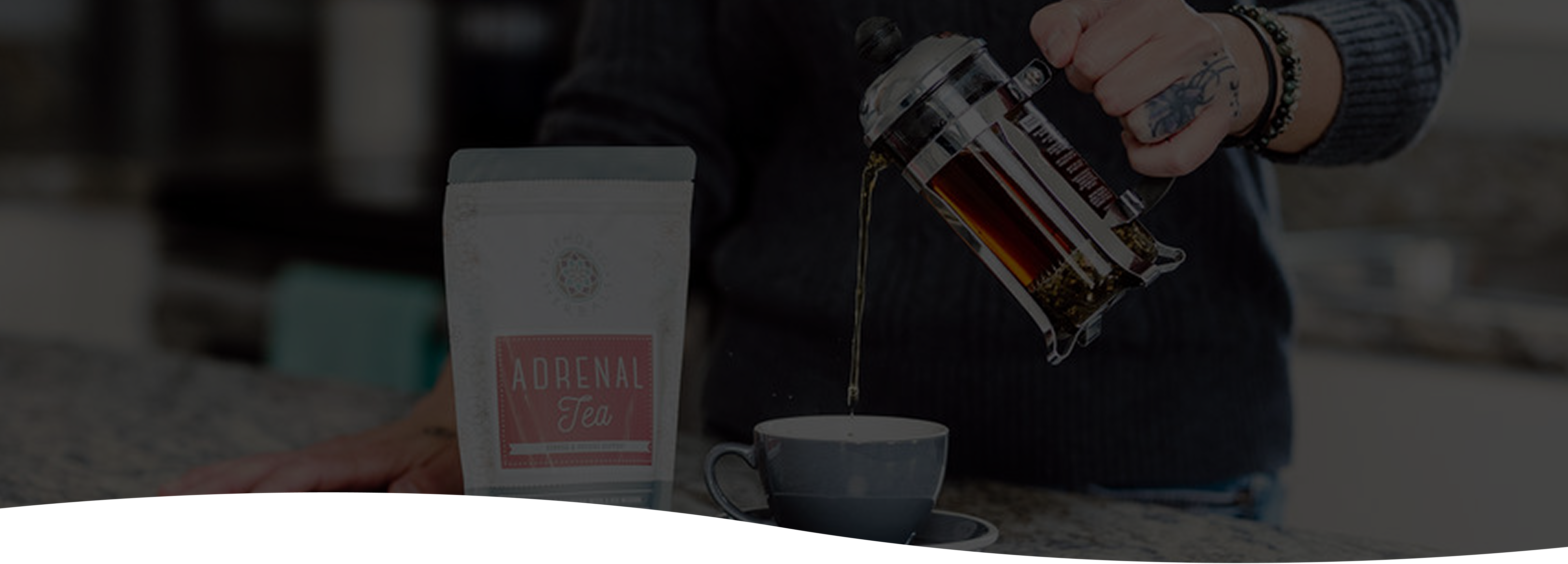 Person pouring tea from a French press into a cup with a bag of herbal tea in the foreground.