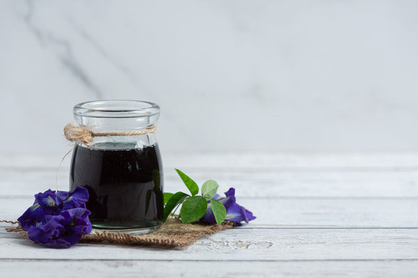 Glass jar filled with dark herbal liquid, tied with twine, placed on a rustic white wooden surface with purple butterfly pea flowers and green leaves beside it.