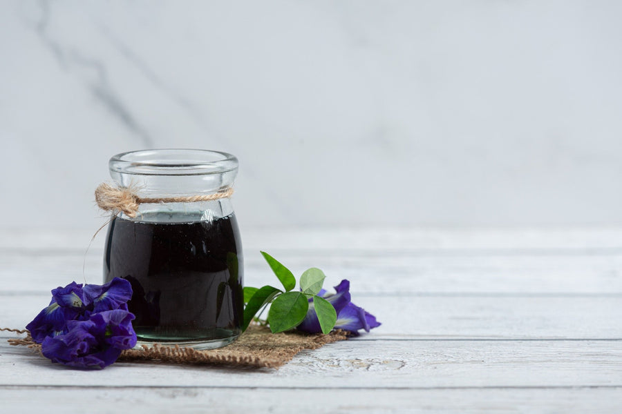 Glass jar filled with dark herbal liquid, tied with twine, placed on a rustic white wooden surface with purple butterfly pea flowers and green leaves beside it.
