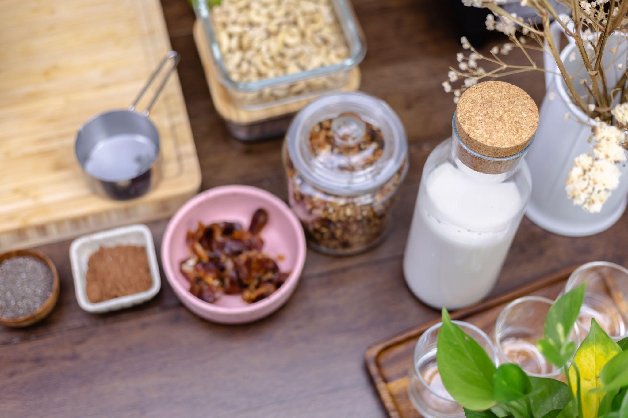 Kitchen countertop with milk, granola, dates, chia seeds, cocoa, measuring cup, and a small green plant.
