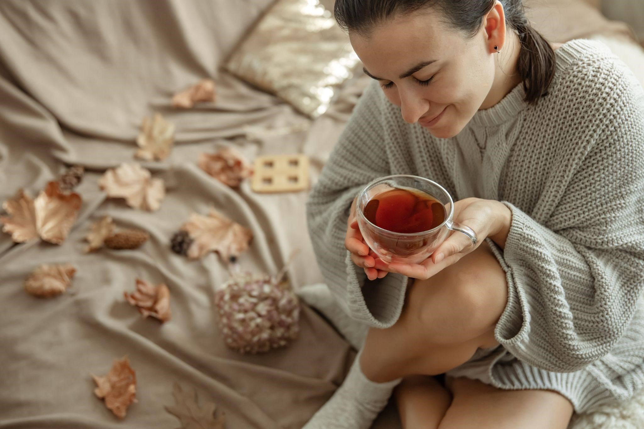 Woman in a cozy sweater sitting on a bed, holding a cup of tea, with autumn leaves and a small flower bouquet scattered around.