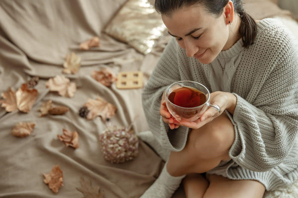 Woman in a cozy sweater sitting on a bed, holding a cup of tea, with autumn leaves and a small flower bouquet scattered around.