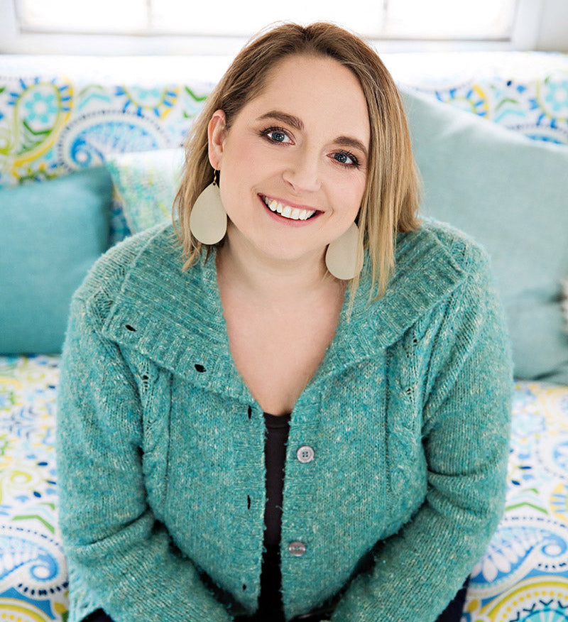 A smiling woman with blonde hair sits on a colorful patterned couch wearing a textured mint green knit cardigan over a black top and dark pants.