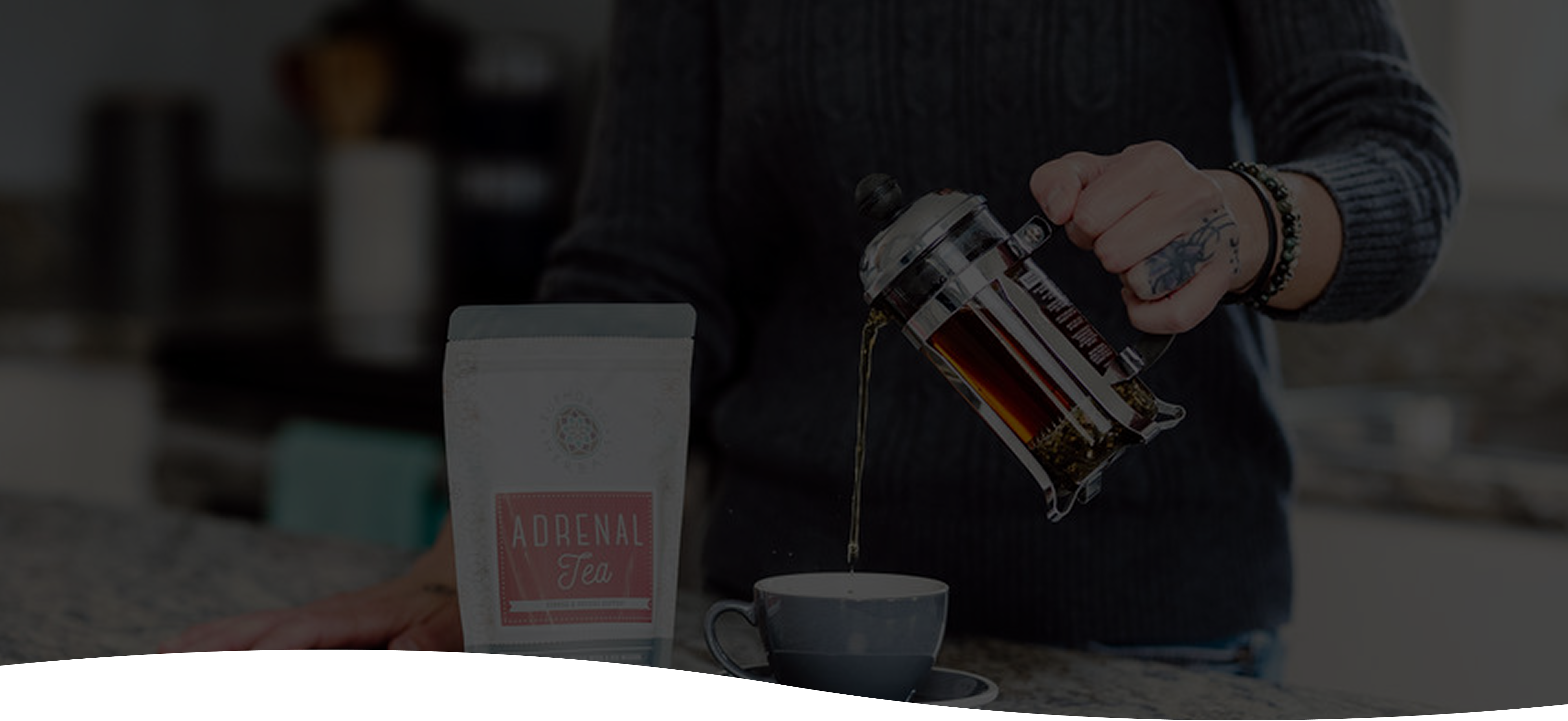 Person pouring tea from a French press into a cup with a bag of herbal tea in the foreground.